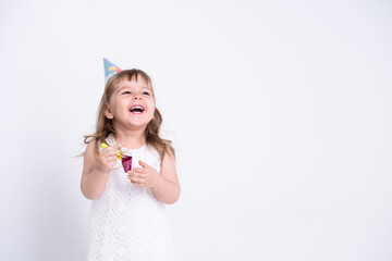 Funny child girl in white dress and birthday hat blowing in whistle on white background