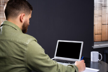 A photo from the back of a man sitting at a writing desk. Fills in documents and works with a laptop with a cup of tea. Remote work at home.
