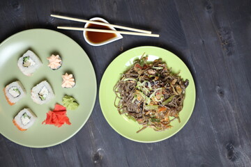 Safood buckwheat noodles stuffes with cucumber and sesame in green plate, rice rolls, white wine glass on black table with chopsticks and soy sauce