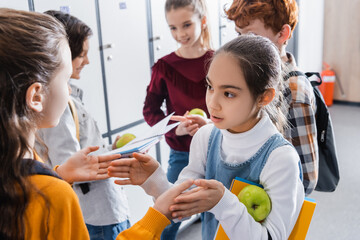 Girls playing patty cake game near classmates with apples and notebooks on blurred background in school