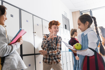 Positive schoolchild talking with schoolgirl holding apple and copybook on blurred foreground in school