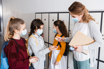 Teacher in protective mask holding sanitizer near pupils in school
