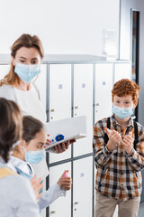 Obraz premium Boy in medical mask holding hand sanitizer near teacher with infrared thermometer and notebook in corridor