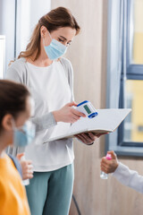 Teacher in medical mask holding infrared thermometer and notebook near pupils on blurred foreground