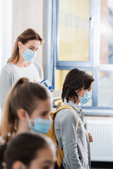 Obraz premium Schoolboy in medical mask standing near teacher with infrared thermometer and classmates