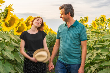 People walking in sunflower field in summer evening at sunset. Portraits of beautiful girl and handsome man. Father and daughter against rural landscape. Weekend trip to countryside, summer holidays