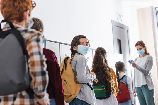Boy in medical mask standing in queue near classmates and teacher with infrared thermometer on blurred background