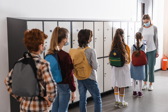 Schoolkids in medical masks standing in queue near teacher with infrared thermometer