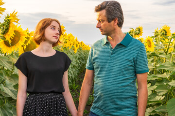 People walking in sunflower field in summer evening at sunset. Portraits of beautiful girl and handsome man. Father and daughter against rural landscape. Weekend trip to countryside, summer holidays