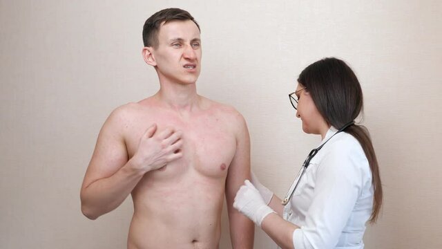 Careful female doctor in white latex gloves and robe examines irritated bare chest of man patient near beige wall in hospital office closeup