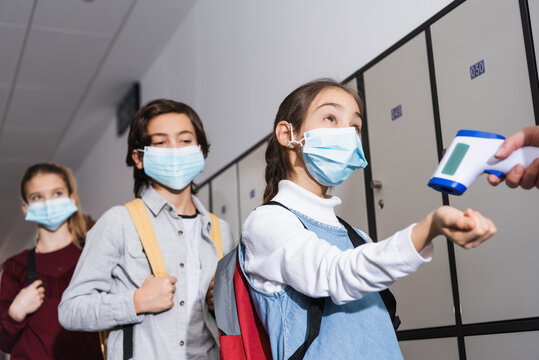 Schoolgirl in medical mask standing near teacher with non contact thermometer and classmates