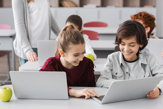 cheerful kids typing on laptop near classmates and teacher on blurred background