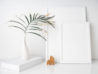 Mock uo poster frames,dry palm leaves  botanical tropical house plant in beautiful white ceramic vase and small wooden house model on white desk and wall background