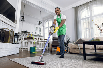 African man removing dust from soft carpet with modern vacuum cleaner. Male cleaner wearing green...