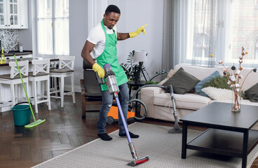 Happy young man in green apron and yellow gloves taking fun while vacuuming soft carpet. Afro american guy enjoying working process during cleaning.