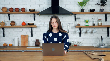Beautiful woman housewife in home kitchen sitting on chair at table work using laptop types on...