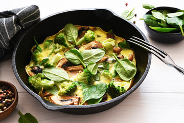 Omelet with spinach and mushrooms in a cast-iron pan on a light wood background. Fritata with spinach.