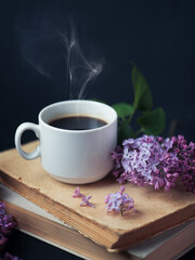 A white cup of hot coffee or tea on a stack of old books, a branch of lilac