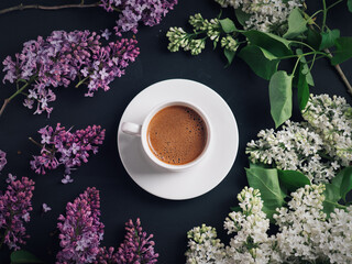 Obraz na płótnie Canvas A cup of homemade coffee top view on a black table with blooming white and purple lilacs