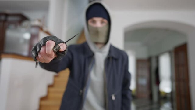 Close-up of knife in male hand with blurred robber standing at background. Masked young Caucasian man robbing wealthy house threatening with cold steel arms. Stealing concept.