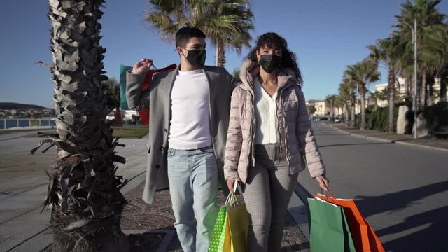 New normal habits and outdoor activity at the Coronavirus pandemic time: young multiracial couple walking and talking each other outside wearing protective mask holding a lot of colored shopping bags