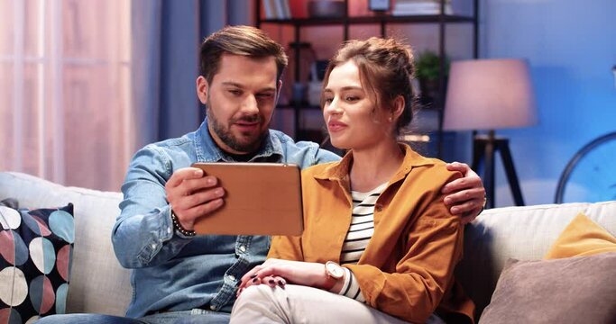 Close up portrait of happy young Caucasian married couple wife and husband videochatting online on tablet sitting on sofa at home and laughing spending evening together watching funny videos on device