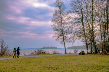 zwei Frauen gehen am Ufer eines Sees mit einzelnen Bäumen bei rosa bewölkter Abendstimmung im Frühjahr spazieren
