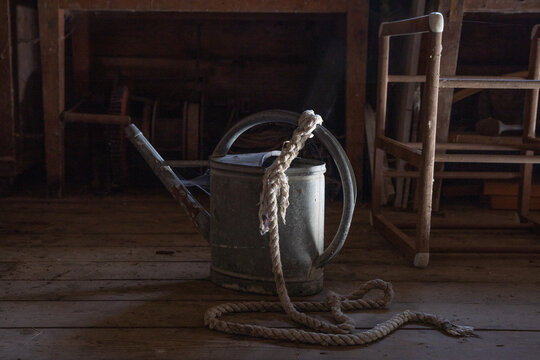 alte, große Giesskanne aus Zink mit angeknotetem dicken Seil in einem Holzschuppen, bereit zum Wasser holen aus einem See