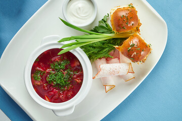 A dish of Ukrainian cuisine - rich pork borsch with Pampushka, green onions and (lard) bacon in a white plate on a blue tablecloth