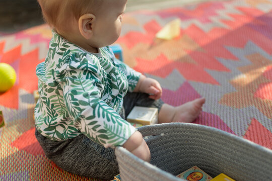 Young Baby Sitting Up On Colorful Outdoor Rug Playing With A Basket Of Blocks