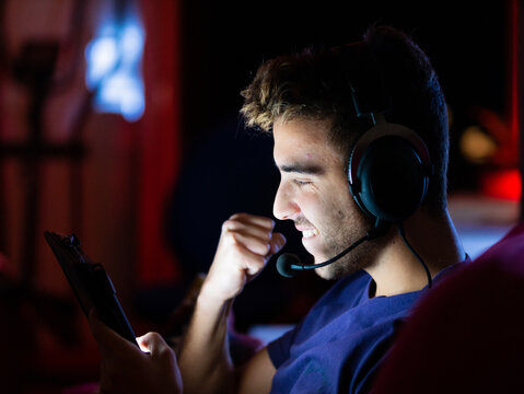 Side view joyful male in headphones browsing modern tablet and raising fist in celebration while sitting in dark room