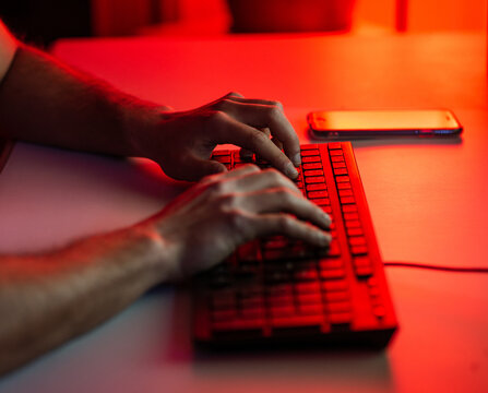 Crop anonymous male typing on keyboard while working on computer and sitting at desk with smartphone in dark room