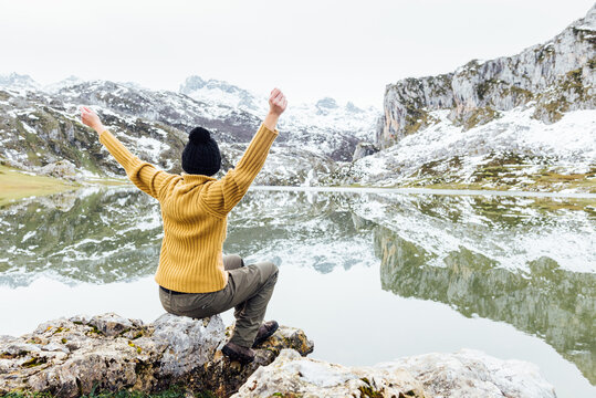 Back view anonymous female wearing warm sweater and black hat sitting with arms outstretched on rough stone on tranquil lakeside and admiring breathtaking landscape of snowy rocky mountains