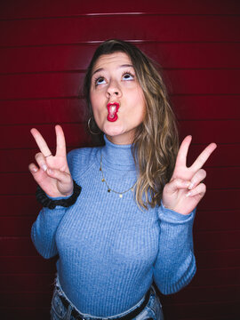 Young female doing grimace showing peace gesture while standing against red striped background