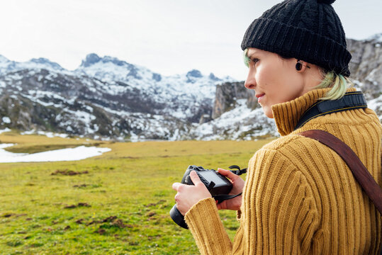 Side view focused young female photographer in warm sweater taking pictures on professional photo camera of majestic rough mountains on grassy highland on clear autumn day