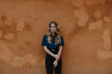 Young mindful blonde female with closed eyes listening to song in wireless headset on brown background in city