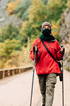 Female hiker in warm clothes with photo camera practicing Nordic walk in autumn forest and looking at camera dreamily