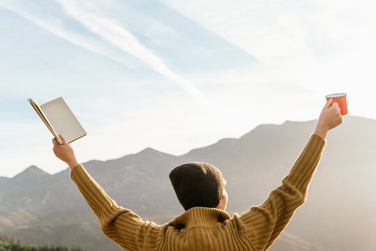Back view of unrecognizable female explorer standing with book and cup of hot drink in raised hands and enjoying freedom in mountains on sunny day
