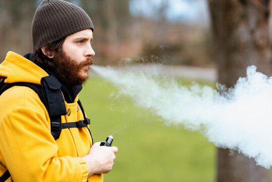 Side view of unshaven male traveler in outerwear with e liquid bottle vaping and looking forward on meadow