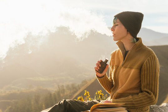 Side view of peaceful female traveler sitting on rock while smoking vape and exhaling cloud of smoke during vacation in mountains