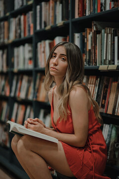 Side view of young unemotional female in sundress sitting with textbook and looking at camera in bookshop