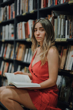 Side view of young female in red sundress with open textbook sitting in bookshop and looking at camera