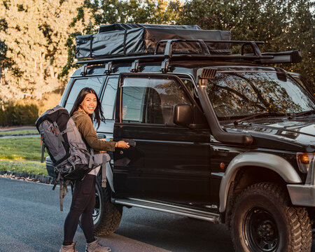 Side view of Asian female backpacker opening door of offroad automobile during travel in Australia and looking at camera