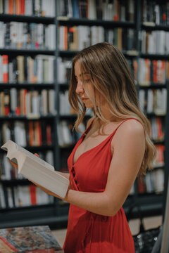 Side view of young female in red sundress with open textbook standing in bookshop