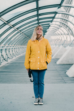 Cheerful female in yellow coat and with umbrella standing under glass roof of tunnel in city and looking at camera