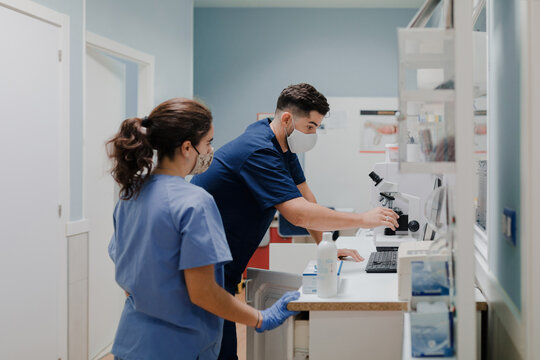 Side view of crop anonymous male medic in uniform and mask using microscope while working in lab near woman colleague