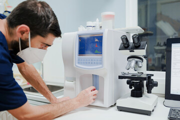 Side view of crop anonymous male doctor in respiratory mask using professional equipment with diagrams on display near microscope in lab