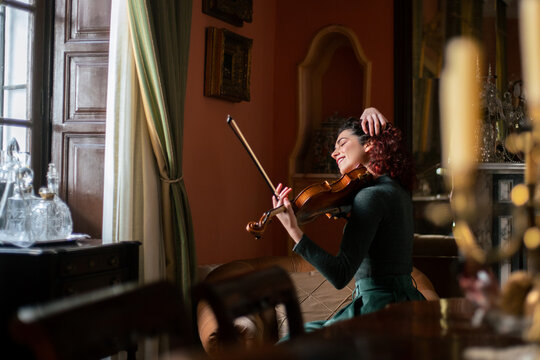 Side view of cheerful female musician touching hair while sitting on armchair in vintage styled room during violin rehearsal