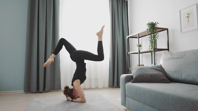 Attractive young woman in black sport clothes doing handstand exercises on floor at home. Concept of yoga, balance and workout.