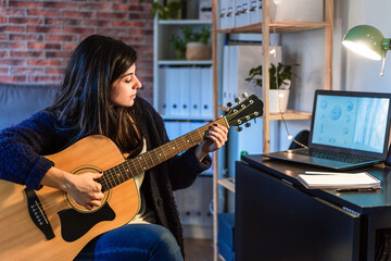 Side view of female playing acoustic guitar while composing music near table with laptop in room with brick wall during remote work